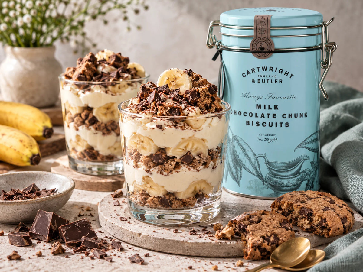 Vanillecreme im Glas mit Banane und Schoko-Biscuit-Crunch, daneben die originale Cartwright-&-Butler-Dose der Milk Chocolate Chunk Biscuits