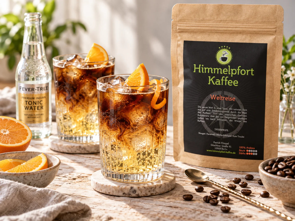 Alt-Text: Frischer Kaffee-Tonic mit Eiswürfeln und Orangenscheibe in einem Glas, daneben die Kaffeetüte „Weltreise“ von Himmelpfort Kaffee sowie Kaffeebohnen und Orangenspalten in einer hellen, natürlichen Food-Szene.