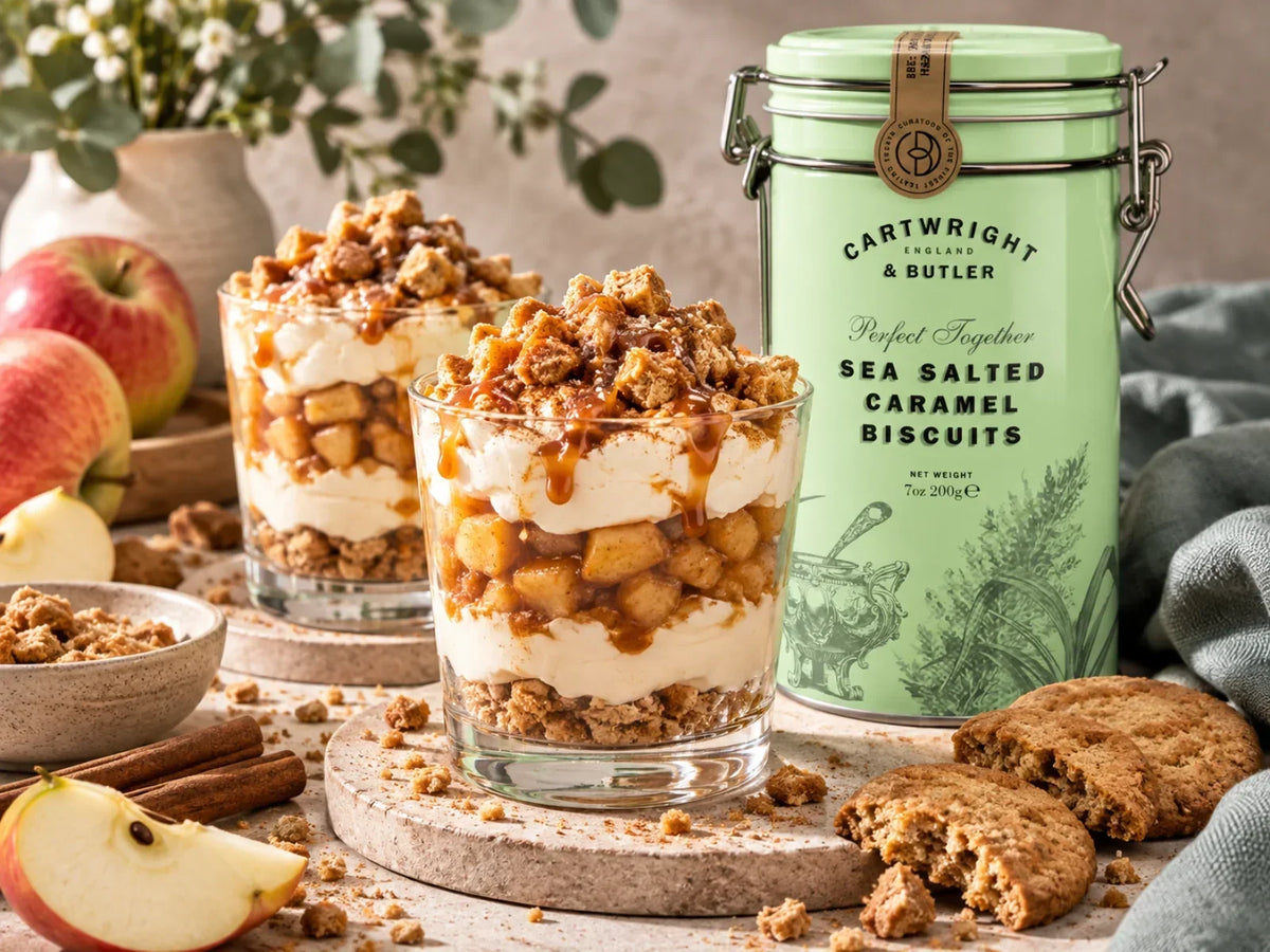 Apfel-Dessert im Glas mit Zimtäpfeln, Vanillecreme und Salted-Caramel-Biscuit-Crunch, daneben die originale Cartwright-&-Butler-Dose der Sea Salted Caramel Biscuits