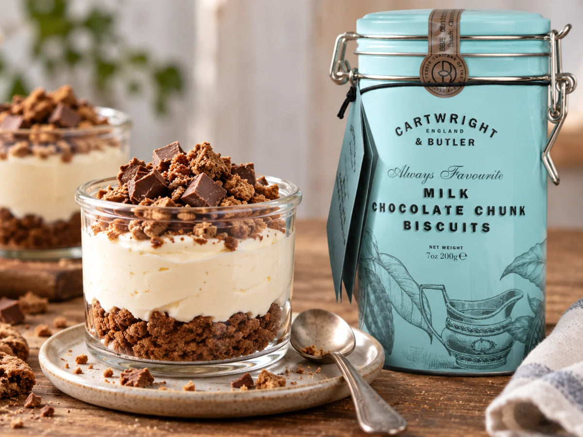 Rustikales Cheesecake-Dessert im Glas mit zerbröselten Schokoladenkeksen, cremiger Füllung und Schokoladenstückchen, daneben die hellblaue Keksdose von Cartwright & Butler.